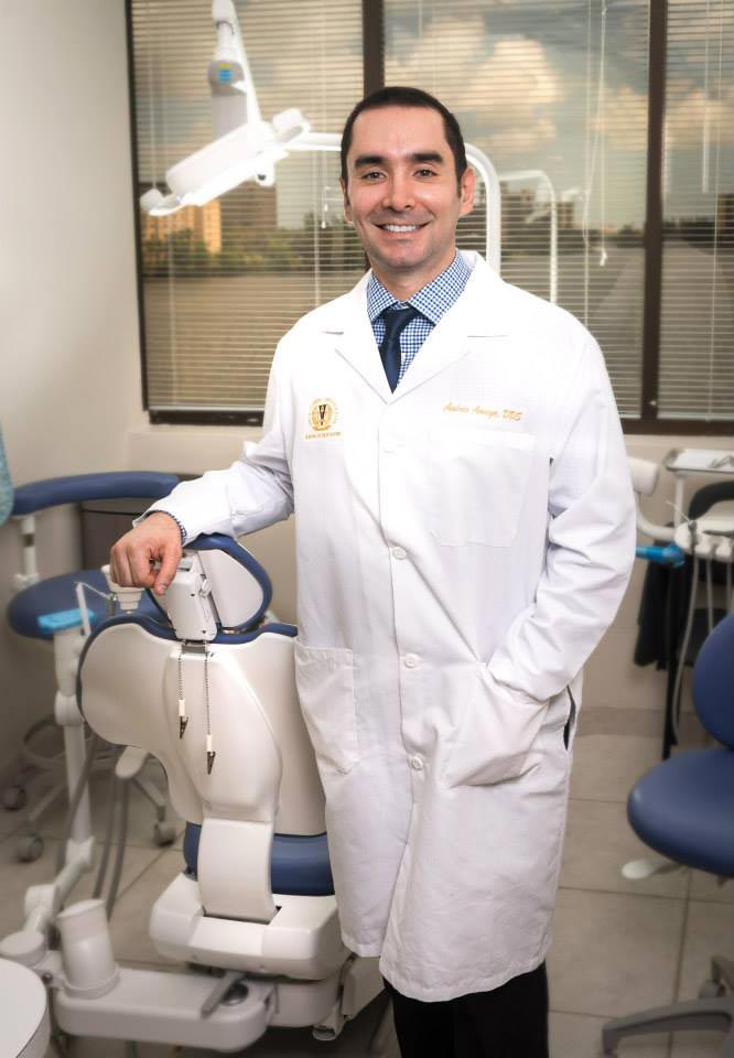 The image shows a man wearing a white lab coat standing next to a dental chair in an office setting, smiling at the camera.