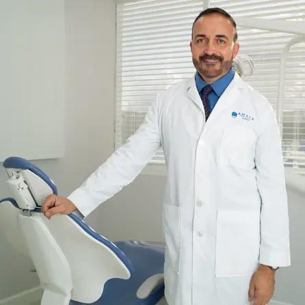 A man wearing a white lab coat stands confidently next to a dental chair in an office setting.