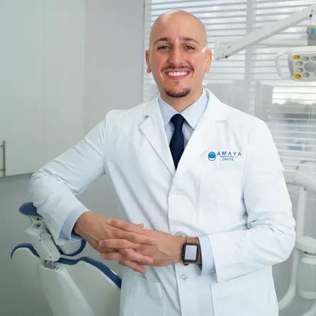 The image shows a smiling dental professional standing in front of a dental chair with a logo on his white coat that reads  AMAYA,  wearing glasses, and posing confidently for the camera.