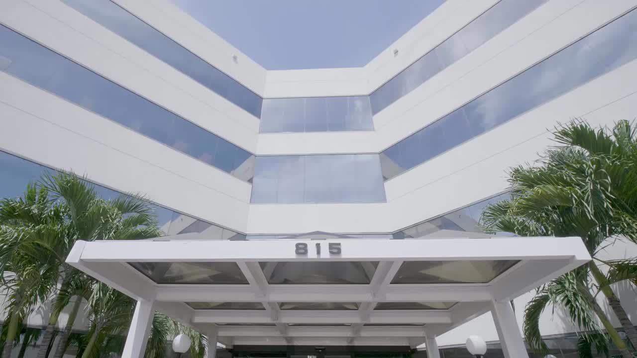 An exterior view of a modern building entrance with a sign displaying  18  above the door, set against a backdrop of palm trees and a clear sky.