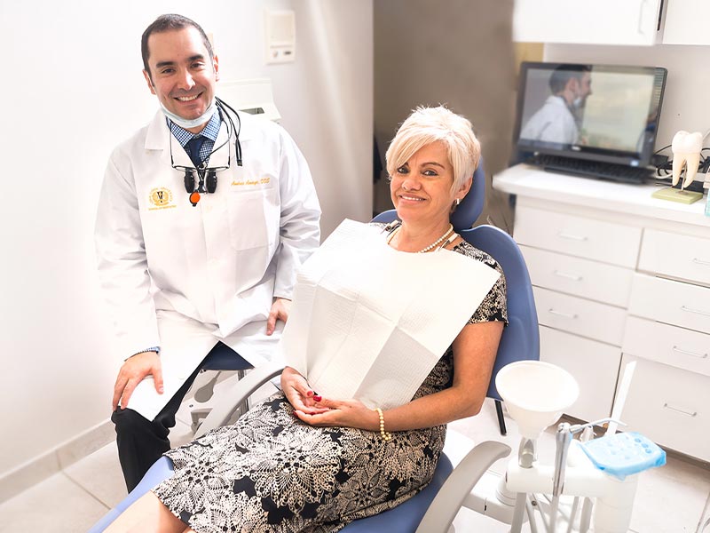 A dentist sitting at a dental chair with a patient, both wearing masks, indicating a medical setting during a pandemic.
