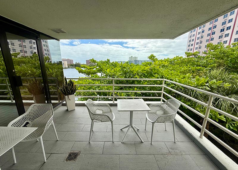 The image shows an outdoor balcony with a view of a body of water and greenery, featuring a railing, chairs, and table.