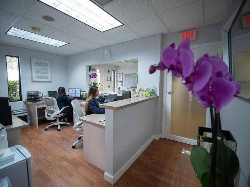 The image shows an interior view of an office space with a reception area, featuring a desk with a computer monitor, a person sitting at the desk, a potted plant, and a purple flower arrangement on the counter.
