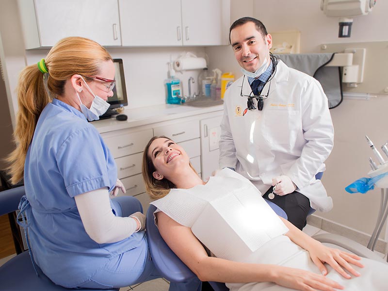 A dentist examining a patient s teeth with a magnifying glass and light, assisted by two dental hygienists, one on each side.