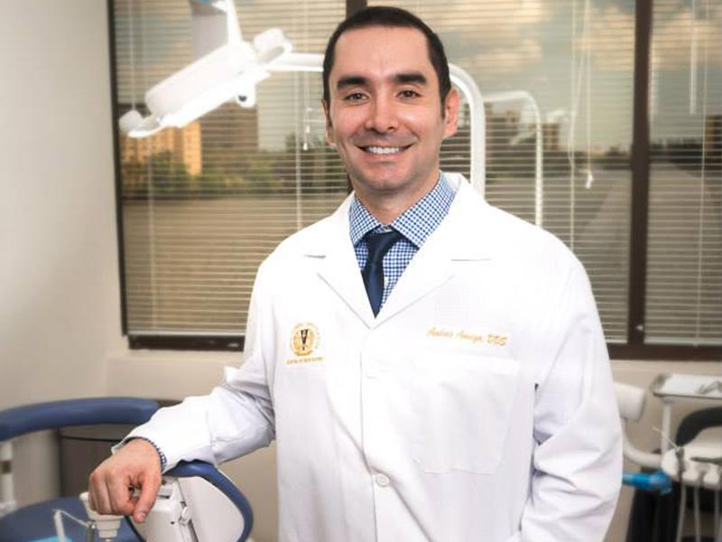 The image shows a man wearing a white lab coat standing in front of a dental chair with dental equipment in the background, smiling at the camera.