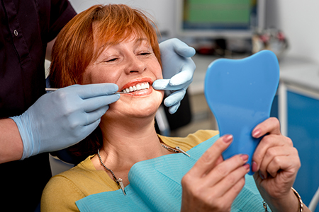 A woman with red hair wearing glasses is sitting in a dental chair, holding up a blue toothbrush, smiling at the camera, surrounded by dental equipment and professionals in a dental office setting.