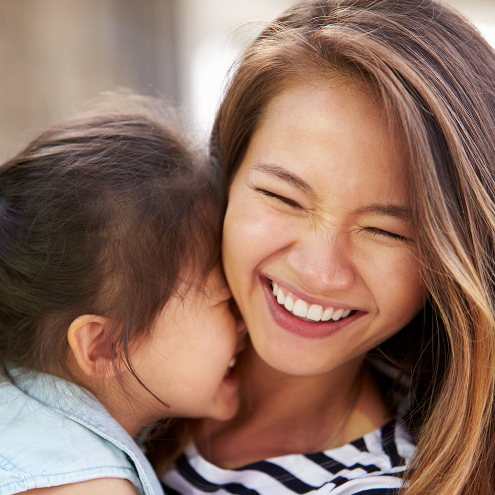 The image shows a woman with a young child, both smiling and looking at each other, with the woman holding the child close to her face.